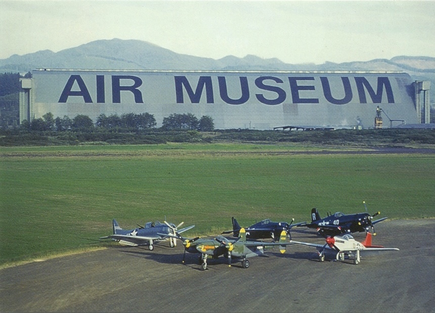 wide photo of a hangar with 'air museum' painted in helvetica on the side with several planes on the runway in the foreground