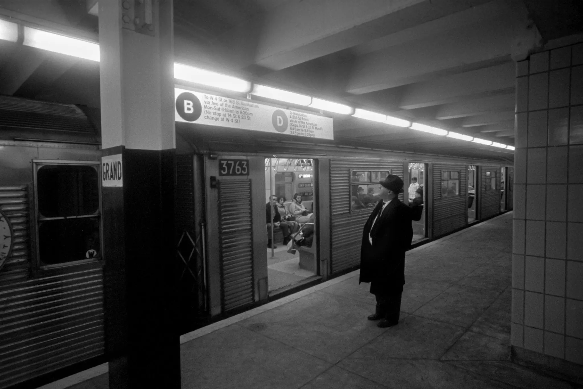 black and white photo of a man standing on the subway platform with a train before him, looking at the new Helvetica subway line signage